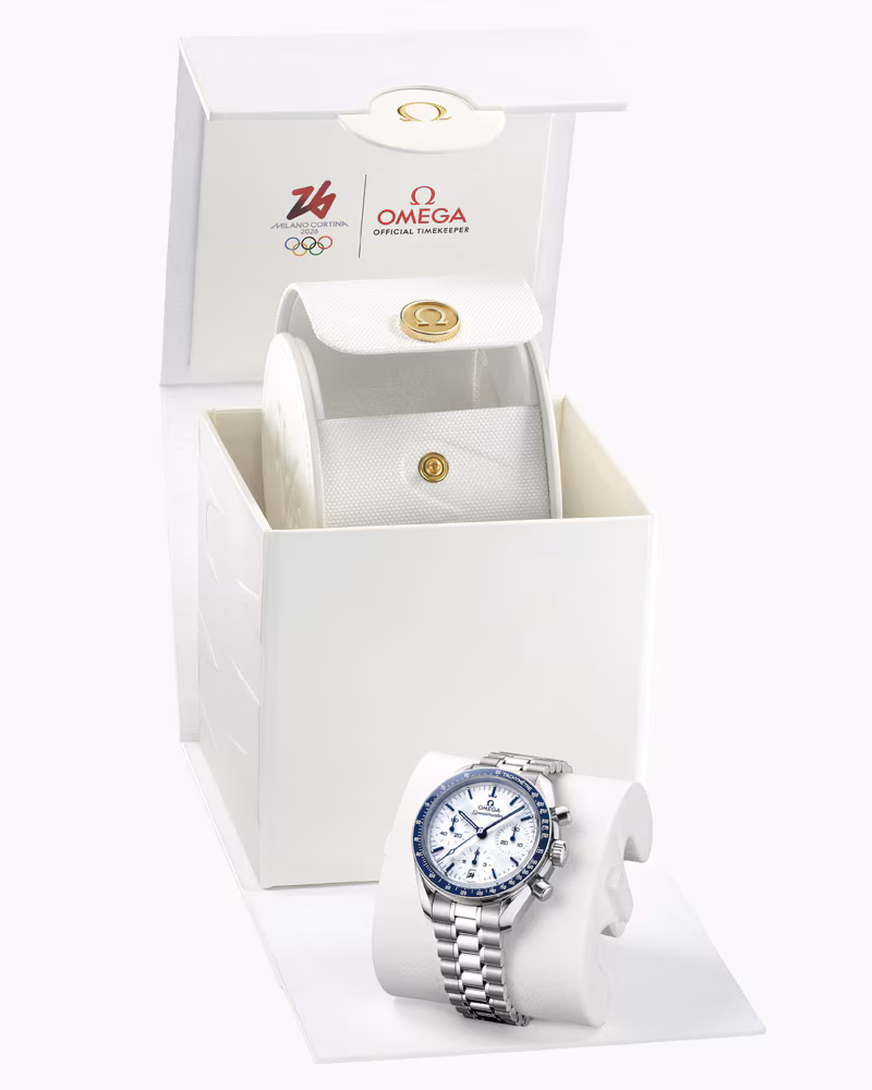 OMEGA Speedmaster Milano Cortina 2026 watch with stainless steel bracelet displayed beside its official white OMEGA Olympic presentation box featuring Milano Cortina 2026 and OMEGA logos.